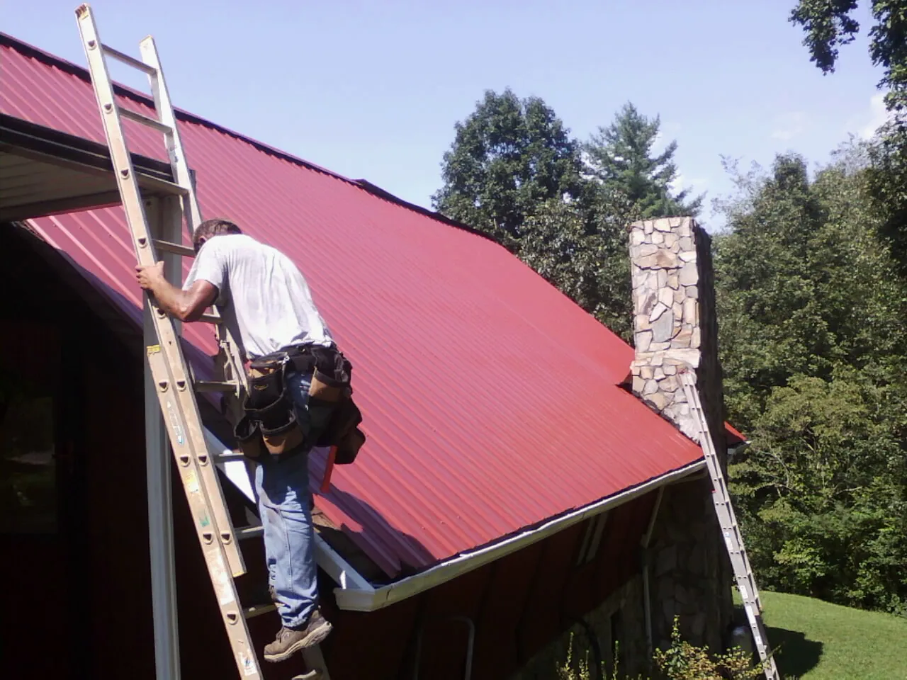 Roofer on a ladder beside a red metal roof and stone chimney — Soffit & Fascia Repair by Roofing Brothers in Gracewood